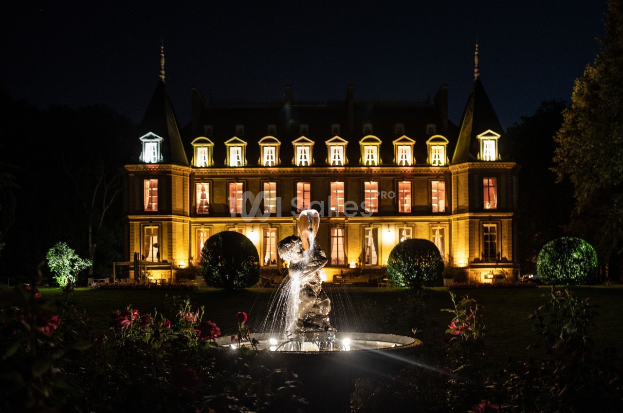 Façade d'un château illuminé la nuit avec une fontaine ornée de statues au premier plan.