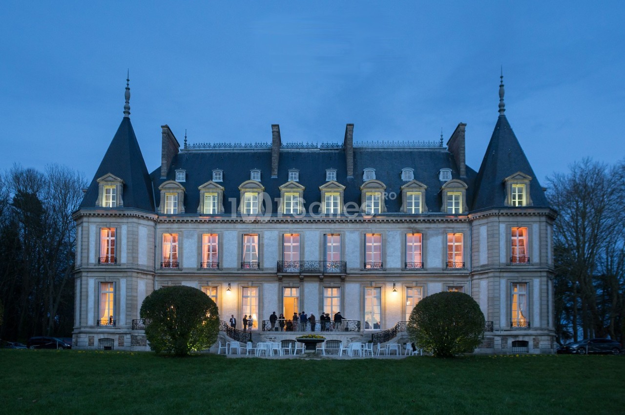 Façade éclairée d'un château au crépuscule, avec des invités rassemblés sur la terrasse et des chaises sur la pelouse.