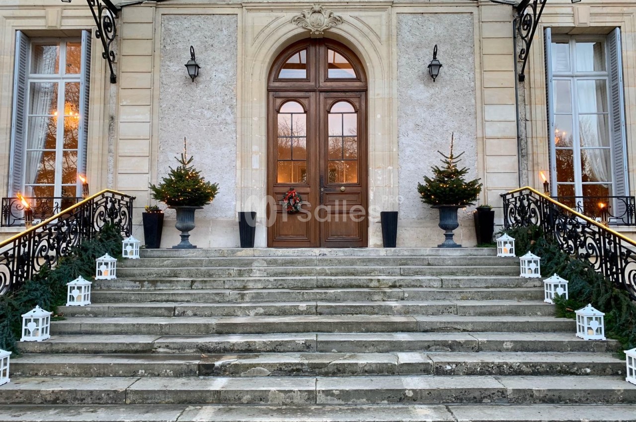 Entrée d'un bâtiment avec porte en bois, décorée de sapins, lanternes blanches et une couronne de Noël.