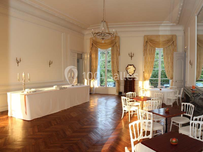 Salle lumineuse avec parquet en bois, tables et chaises blanches, grandes fenêtres et rideaux dorés.