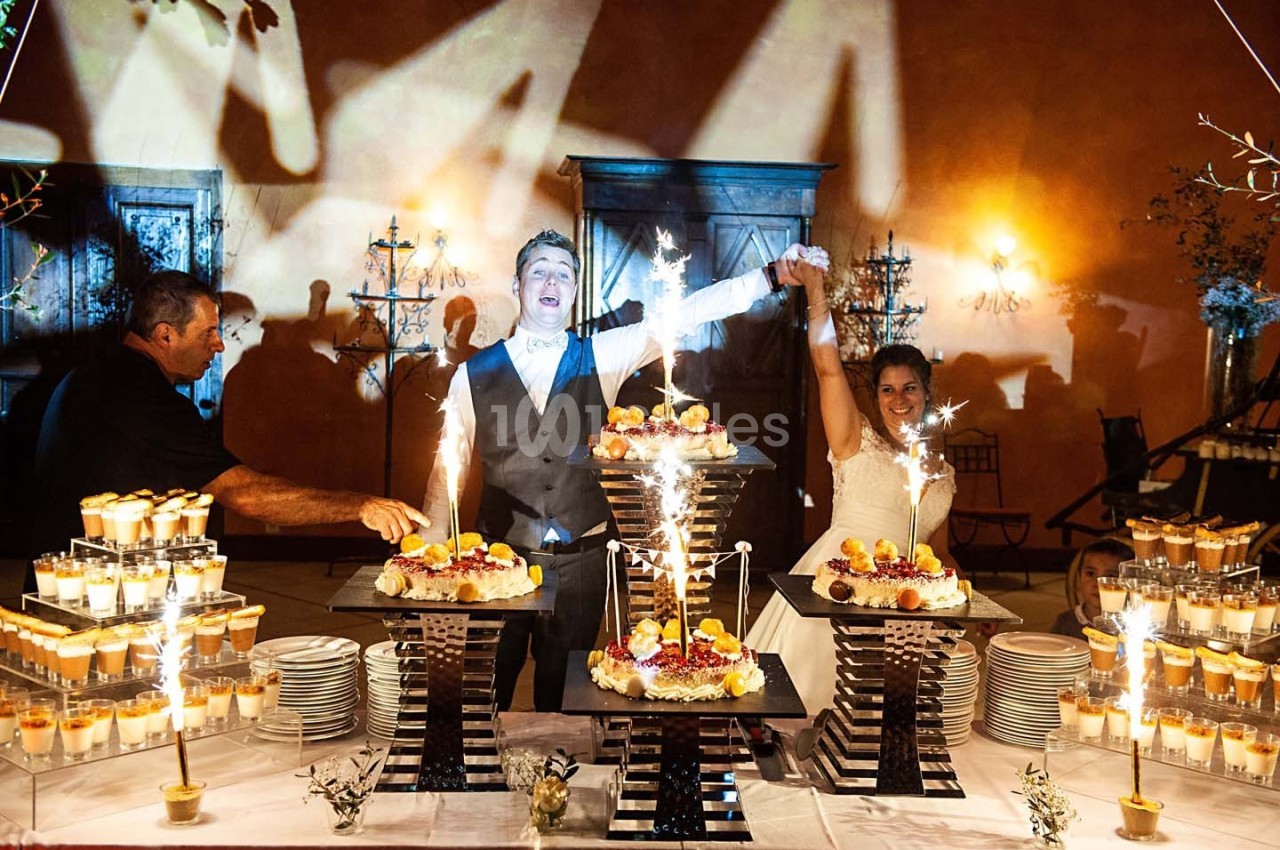 Un couple de mariés souriants coupe un gâteau entouré de desserts et de fontaines lumineuses lors d'une réception.