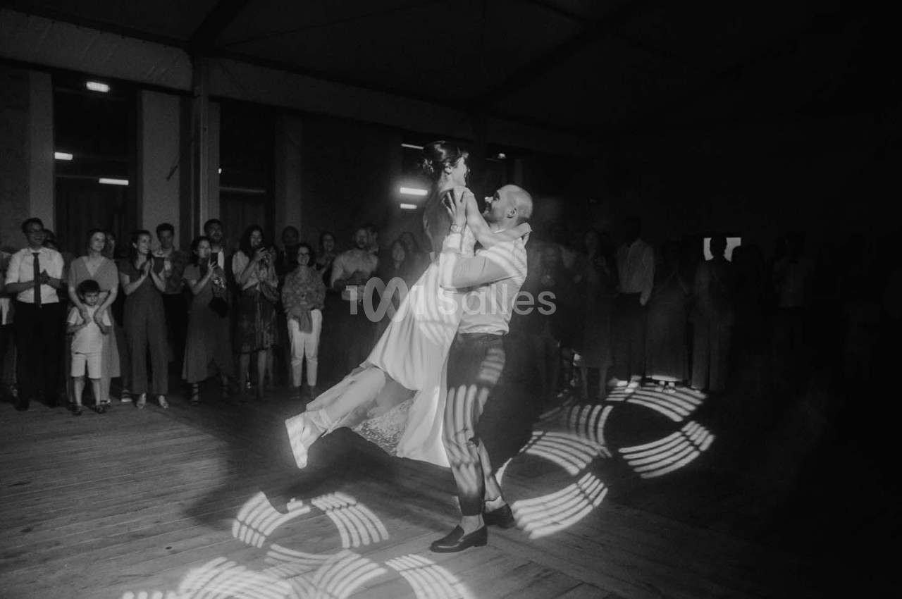 Un couple danse au centre d'une salle sombre, entouré d'invités applaudissant, avec des motifs lumineux au sol.
