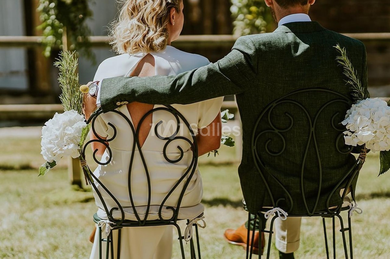 Un couple assis sur des chaises en fer forgé, de dos, lors d'une cérémonie en extérieur par une journée ensoleillée.