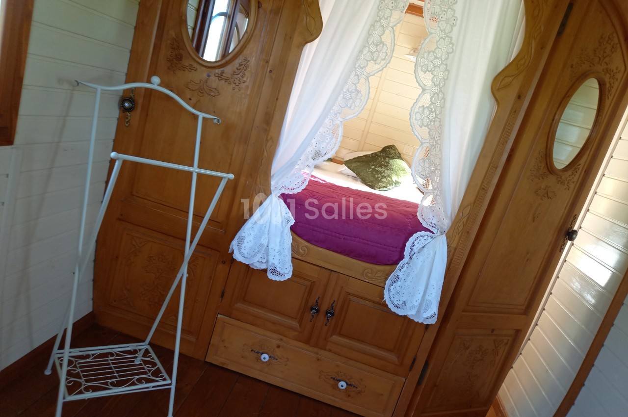 Lit avec draps blancs et coussin vert encadré par des rideaux en dentelle, dans une pièce en bois avec porte-manteau…