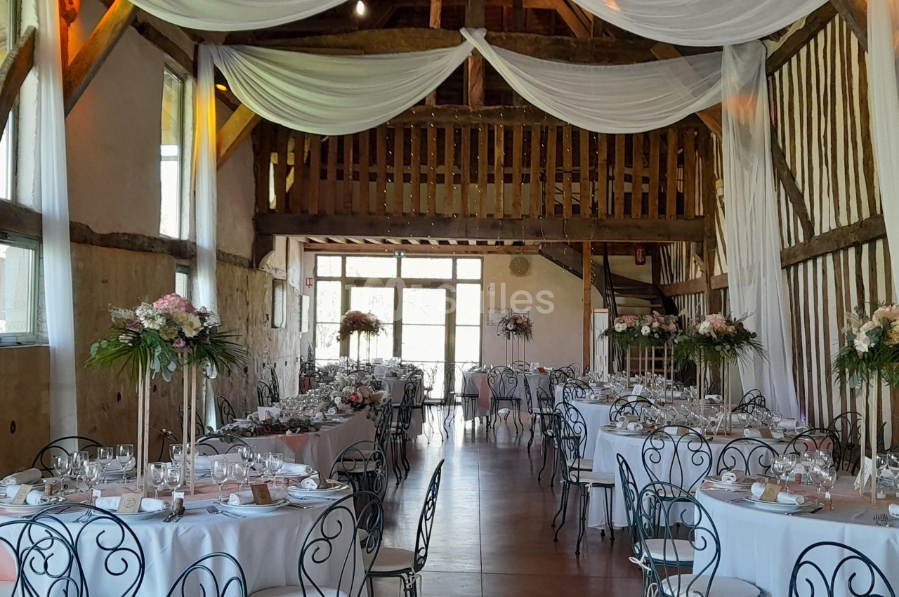 Salle de réception rustique avec tables rondes dressées, chaises en fer forgé et drapés blancs suspendus au plafond.