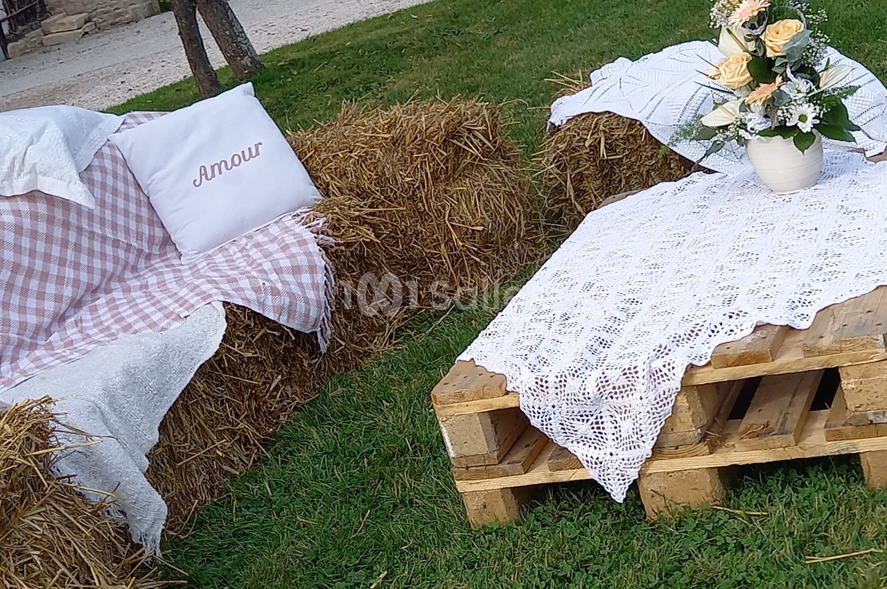 Canapé en bottes de paille avec coussin et nappe à carreaux, table en palettes décorée de dentelle et d'un bouquet de fleurs.