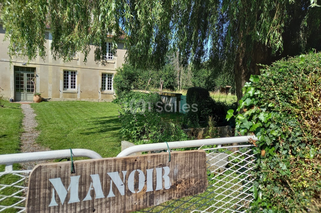 Vue d'un manoir en pierre entouré de verdure, avec une enseigne en bois fixée sur un portail métallique.