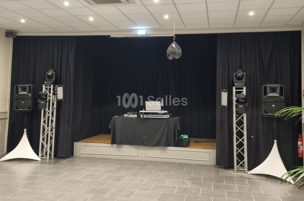 Scène avec rideaux noirs, table de mixage, enceintes sur pieds et éclairage suspendu dans une salle carrelée.