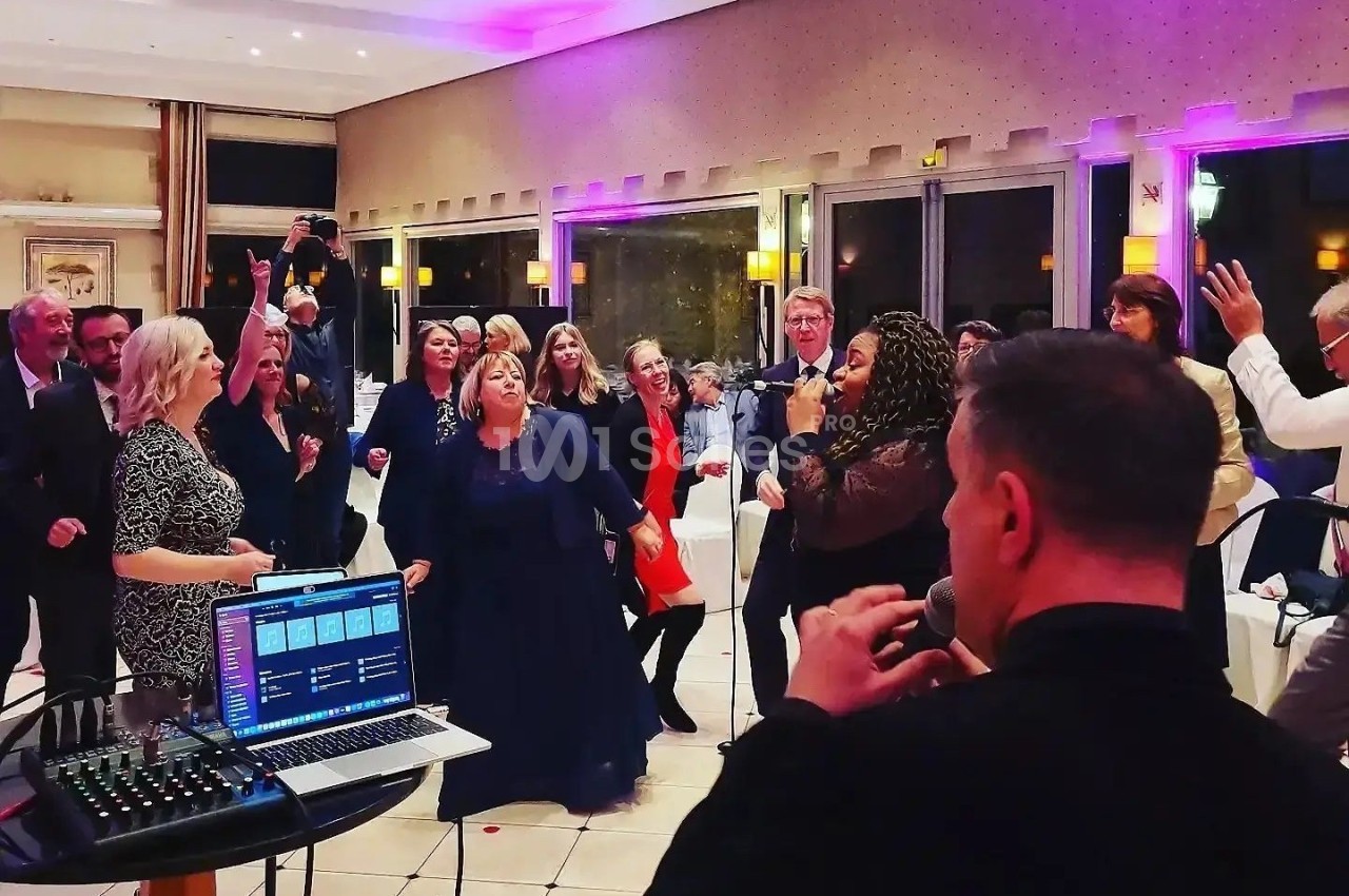 Groupe de personnes dans une salle, certaines dansent tandis qu'une chanteuse et un musicien animent l'événement.