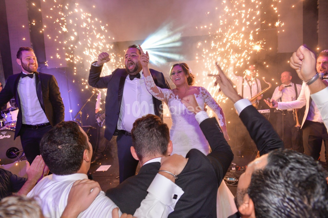 Un couple en tenue de mariage danse entouré d'invités en costume, avec des étincelles et un groupe en arrière-plan.