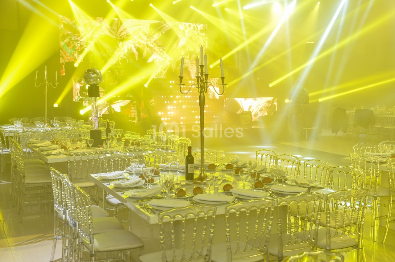 Salle de réception illuminée par des projecteurs jaunes, avec tables dressées et chaises transparentes.