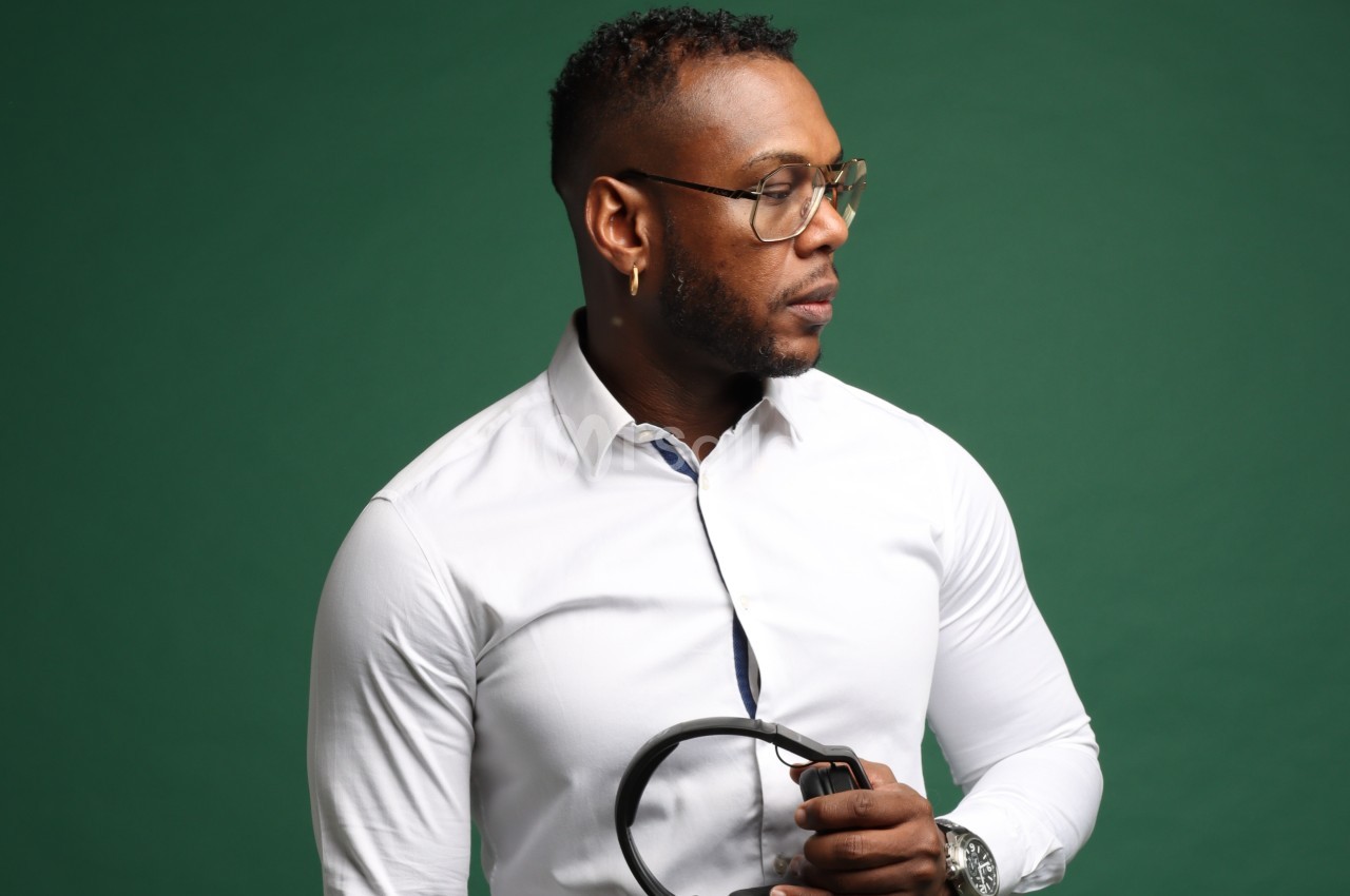 Homme portant une chemise blanche et des lunettes, tenant un casque audio devant un fond vert.