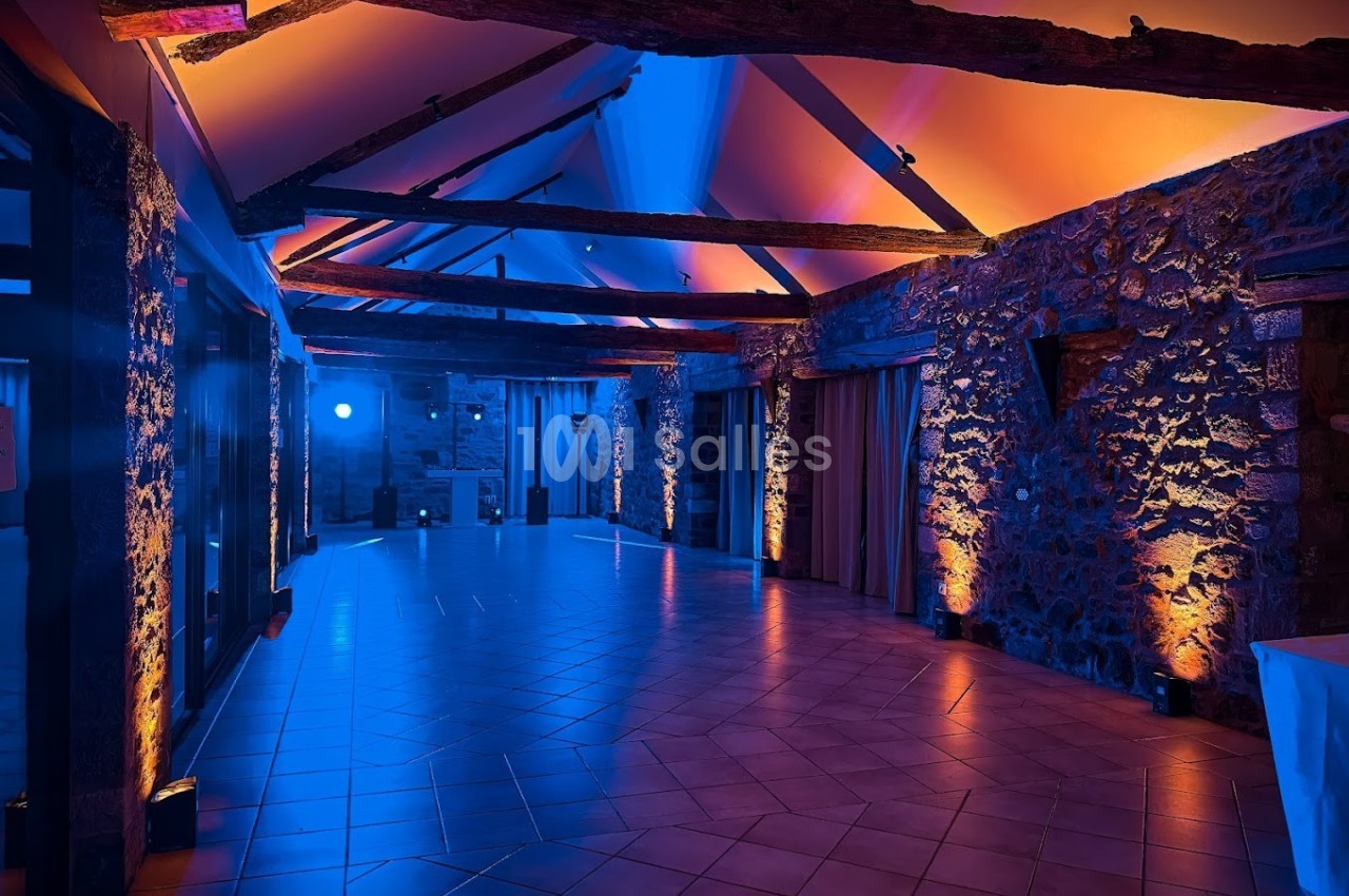 Salle en pierre avec poutres apparentes, éclairée par des lumières colorées bleues et orange, au plafond incliné.
