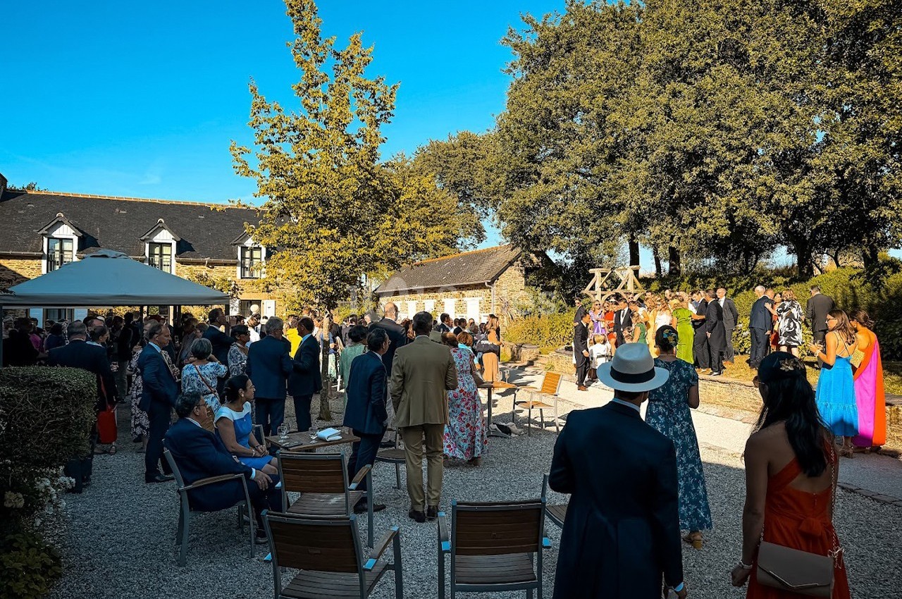 Groupe de personnes rassemblées en extérieur lors d'un événement, près de bâtiments en pierre et d'arbres sous un ciel bleu.