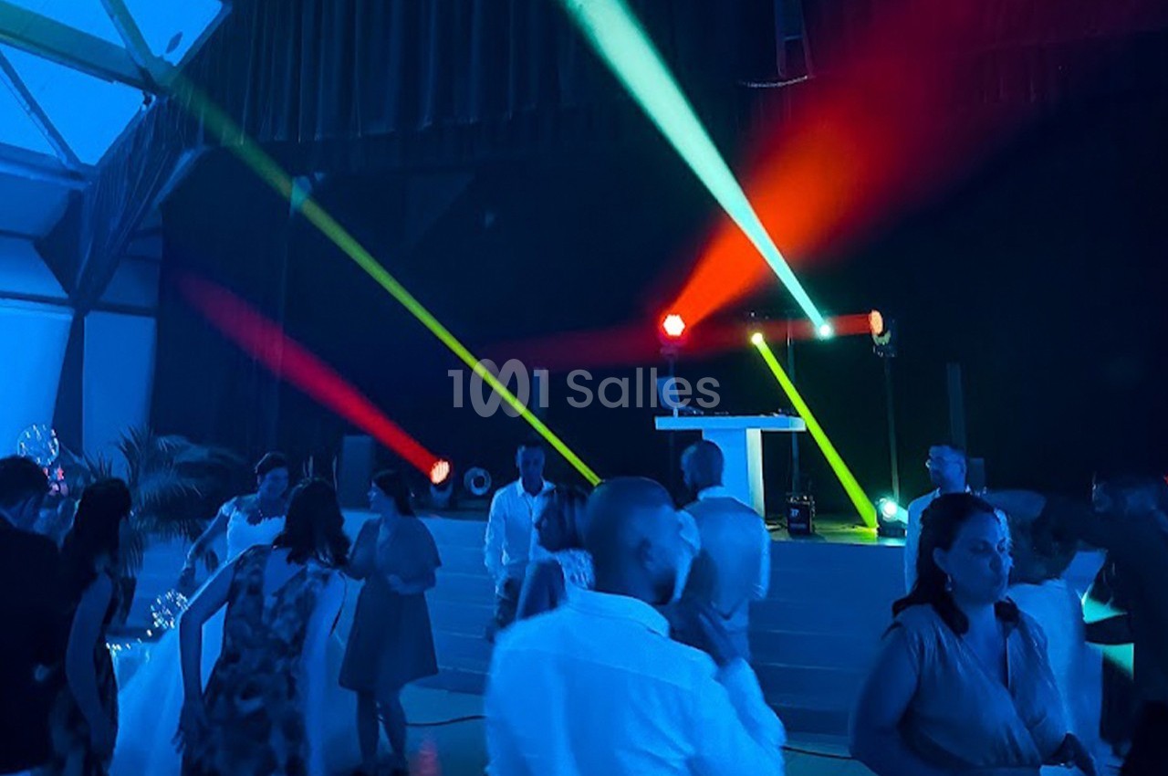 Personnes dansant sous des lumières colorées dans une salle sombre avec une scène équipée de matériel de DJ.