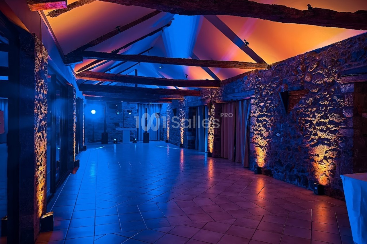 Salle en pierre avec poutres apparentes, éclairée par des lumières colorées bleues et orange, au plafond incliné.