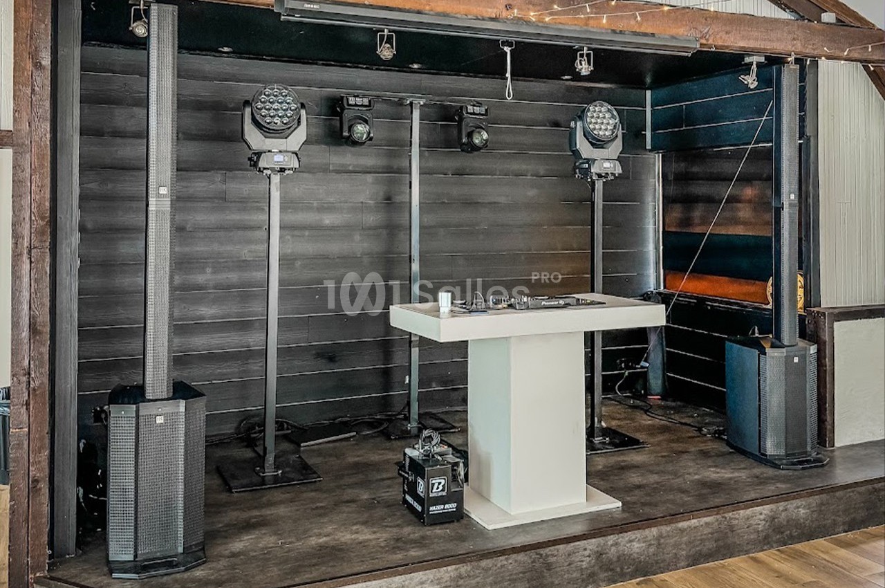 Scène intérieure avec une table de mixage blanche, des projecteurs et des enceintes installés sur une estrade en bois.
