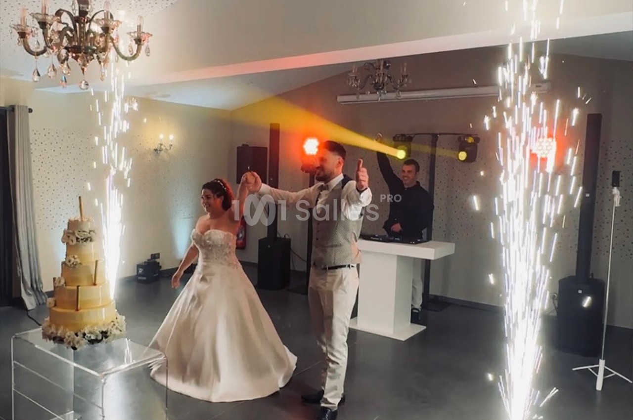 Un couple de mariés danse sous des étincelles près d'un gâteau de mariage dans une salle éclairée.