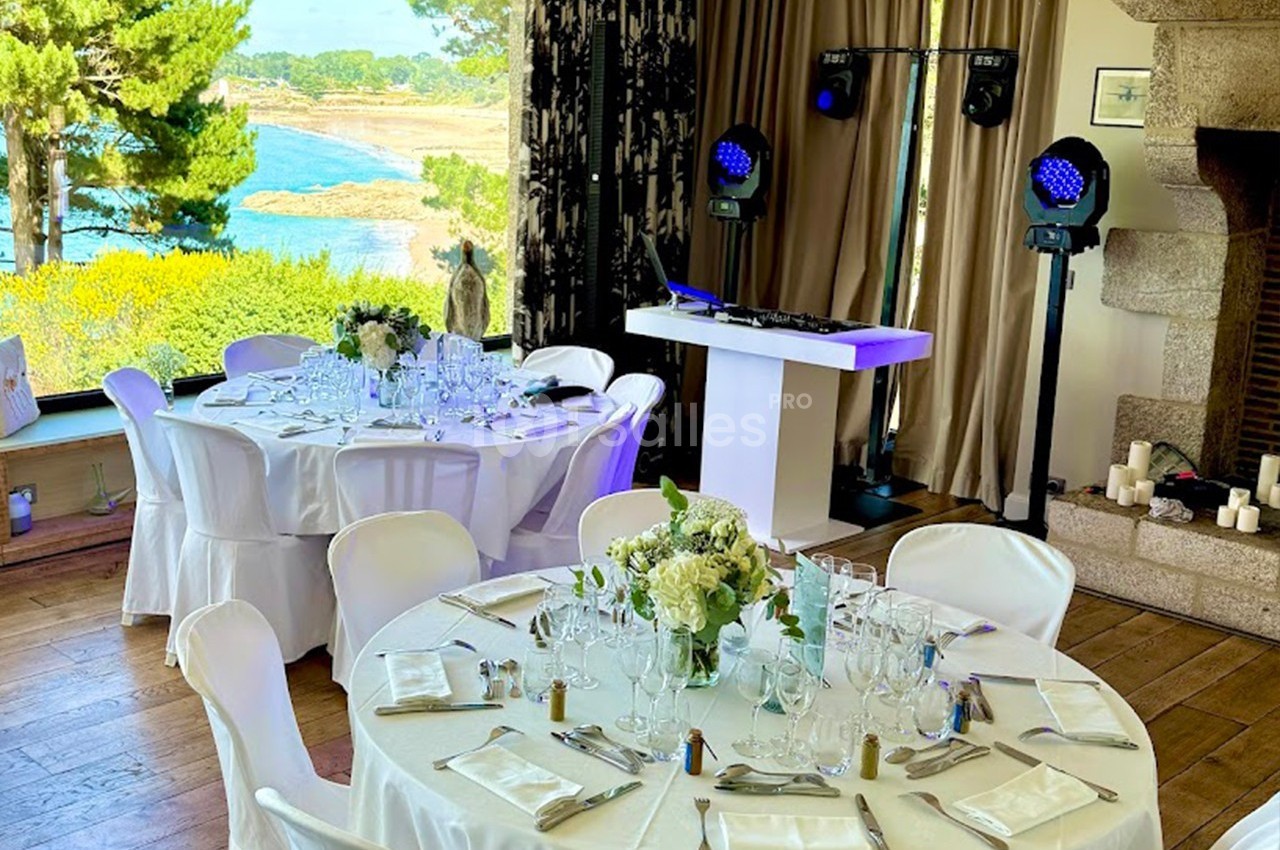 Salle décorée pour un événement avec tables rondes dressées, vue sur un paysage côtier et espace DJ en arrière-plan.