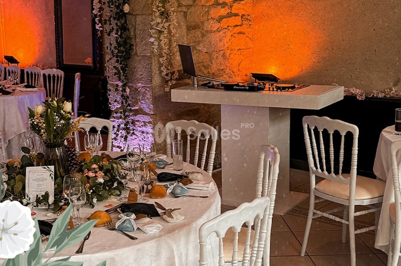 Salle de réception décorée avec des tables rondes dressées, des chaises blanches et une table de DJ en arrière-plan.