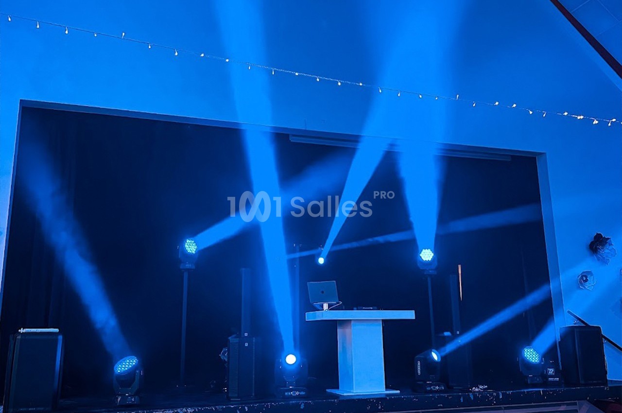 Scène éclairée par des projecteurs bleus avec une table blanche et un ordinateur au centre.