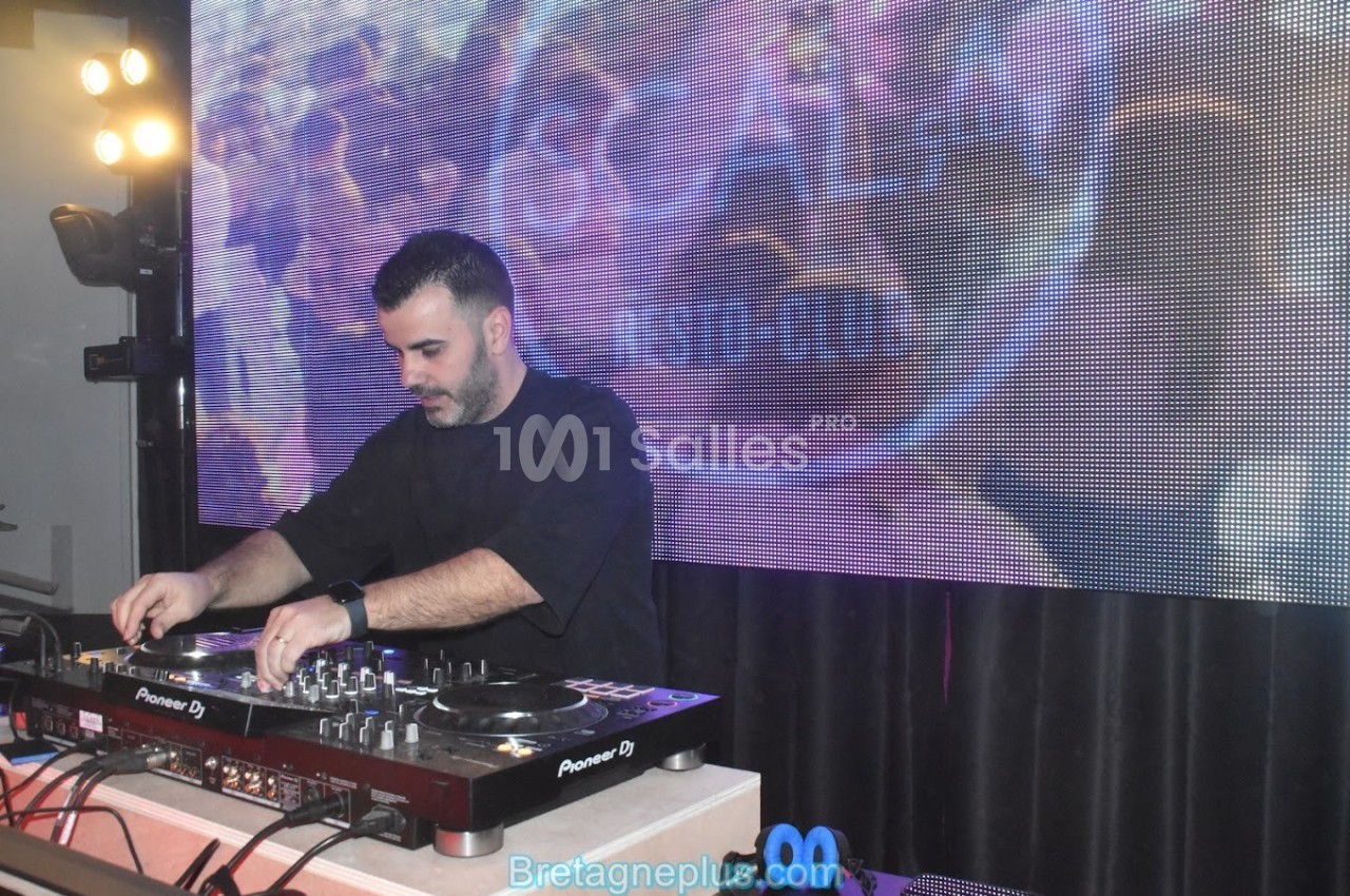 Un DJ mixe de la musique sur une table de mixage devant un écran lumineux affichant des motifs colorés.