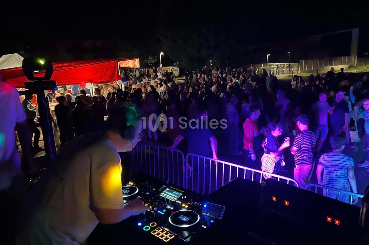 Un DJ mixe sur une scène en plein air face à une foule dansante sous des éclairages colorés, de nuit.