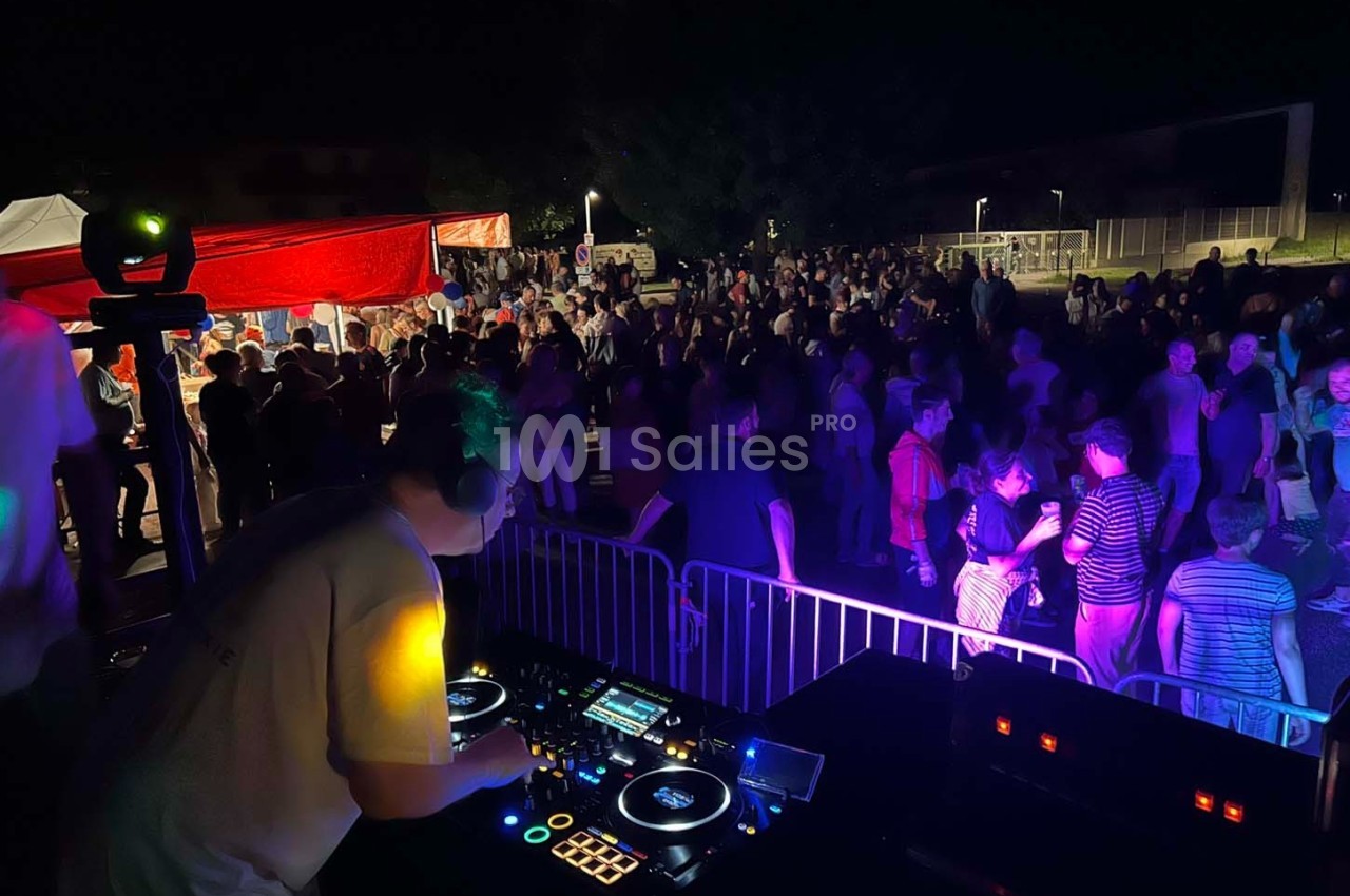 Un DJ mixe sur une scène en plein air face à une foule dansante sous des éclairages colorés, de nuit.