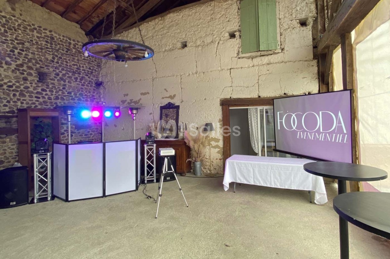 Salle rustique aménagée avec un espace DJ, éclairages colorés, tables hautes et écran affichant ’Foooda Événementiel’.