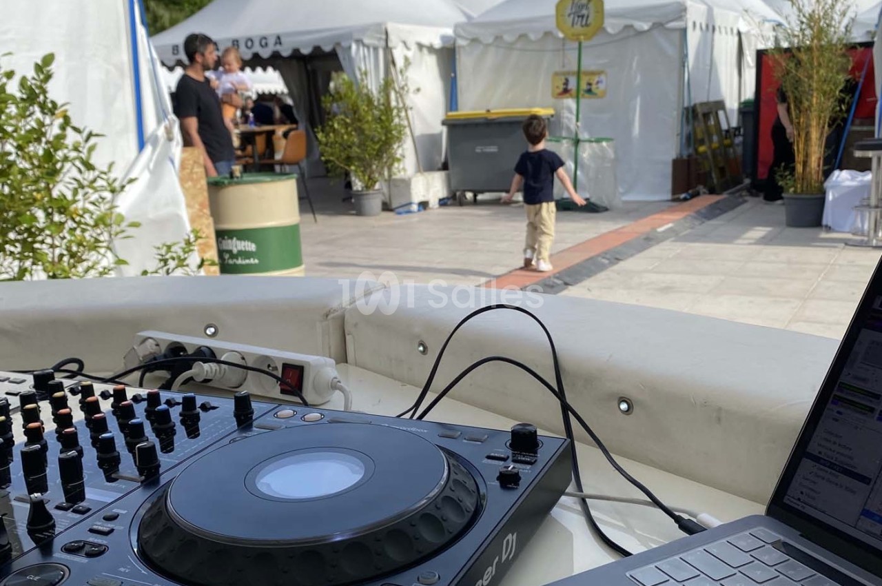 Vue d'une table de mixage et d'un ordinateur portable en extérieur, avec des stands et des personnes en arrière-plan.