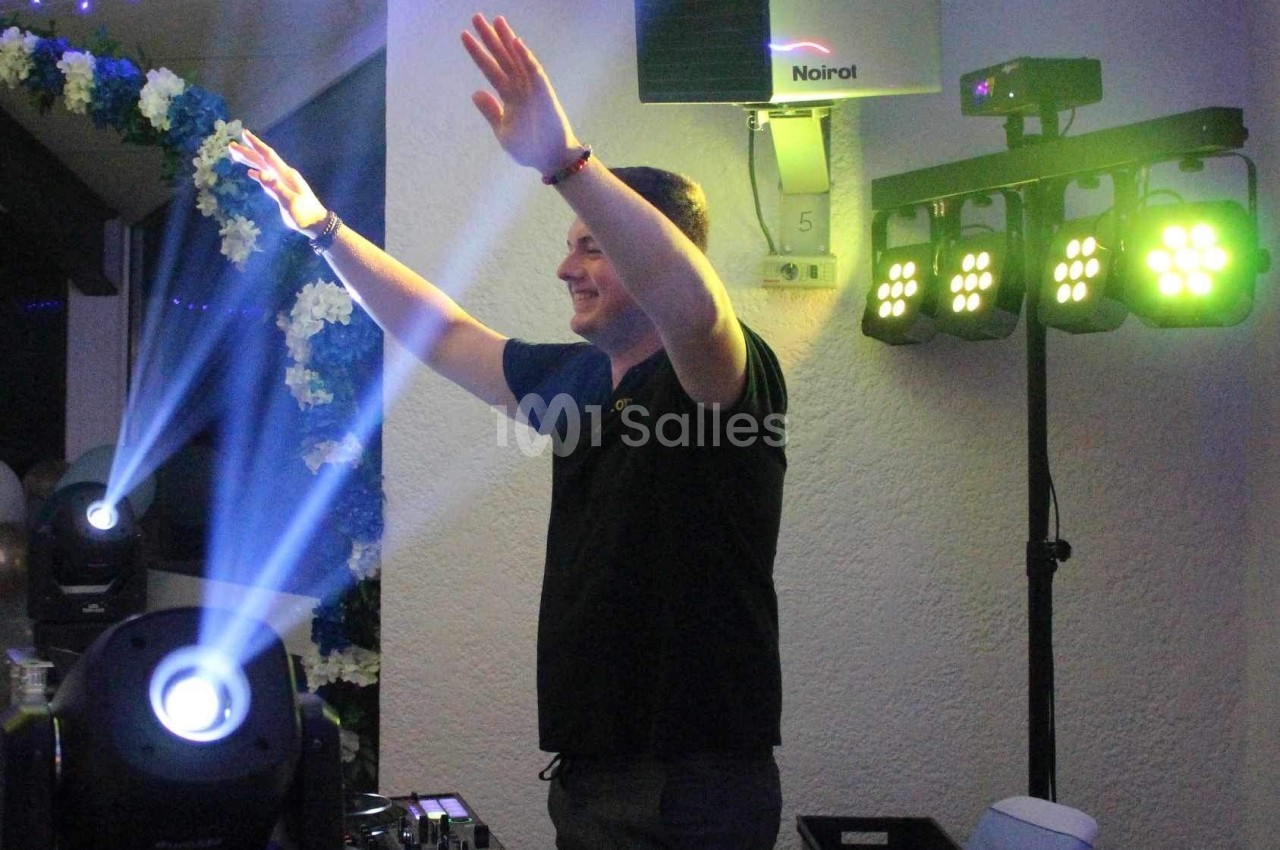 Un DJ anime une soirée avec des jeux de lumières colorés dans une salle décorée de fleurs.