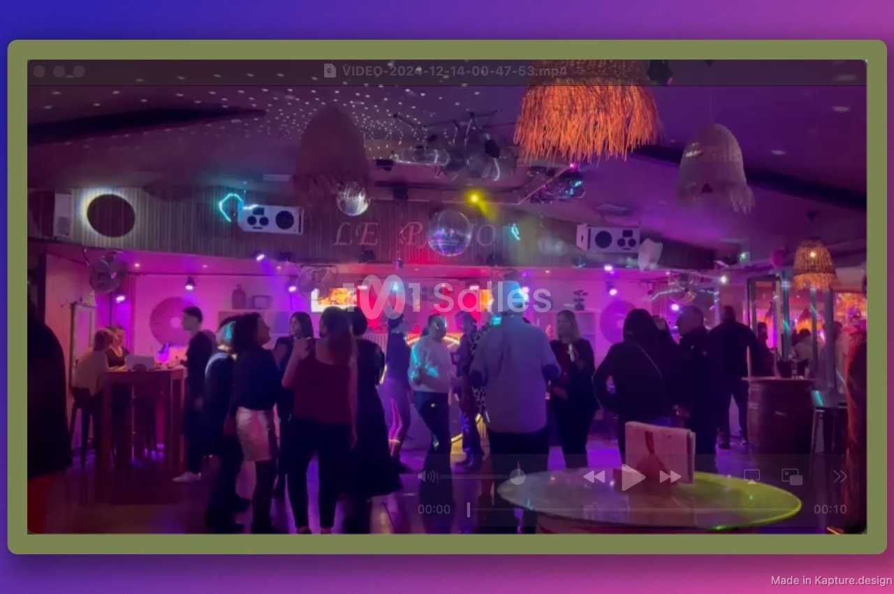 Personnes dansant dans une salle éclairée par des lumières colorées, avec un bar et une décoration festive en arrière-plan.