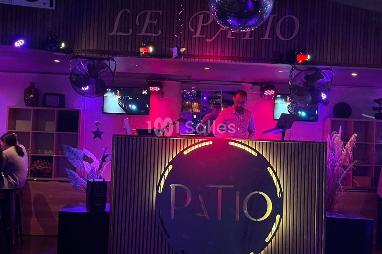 Un DJ mixe sur une scène éclairée par des lumières colorées dans un lieu nommé ’Le Patio’.