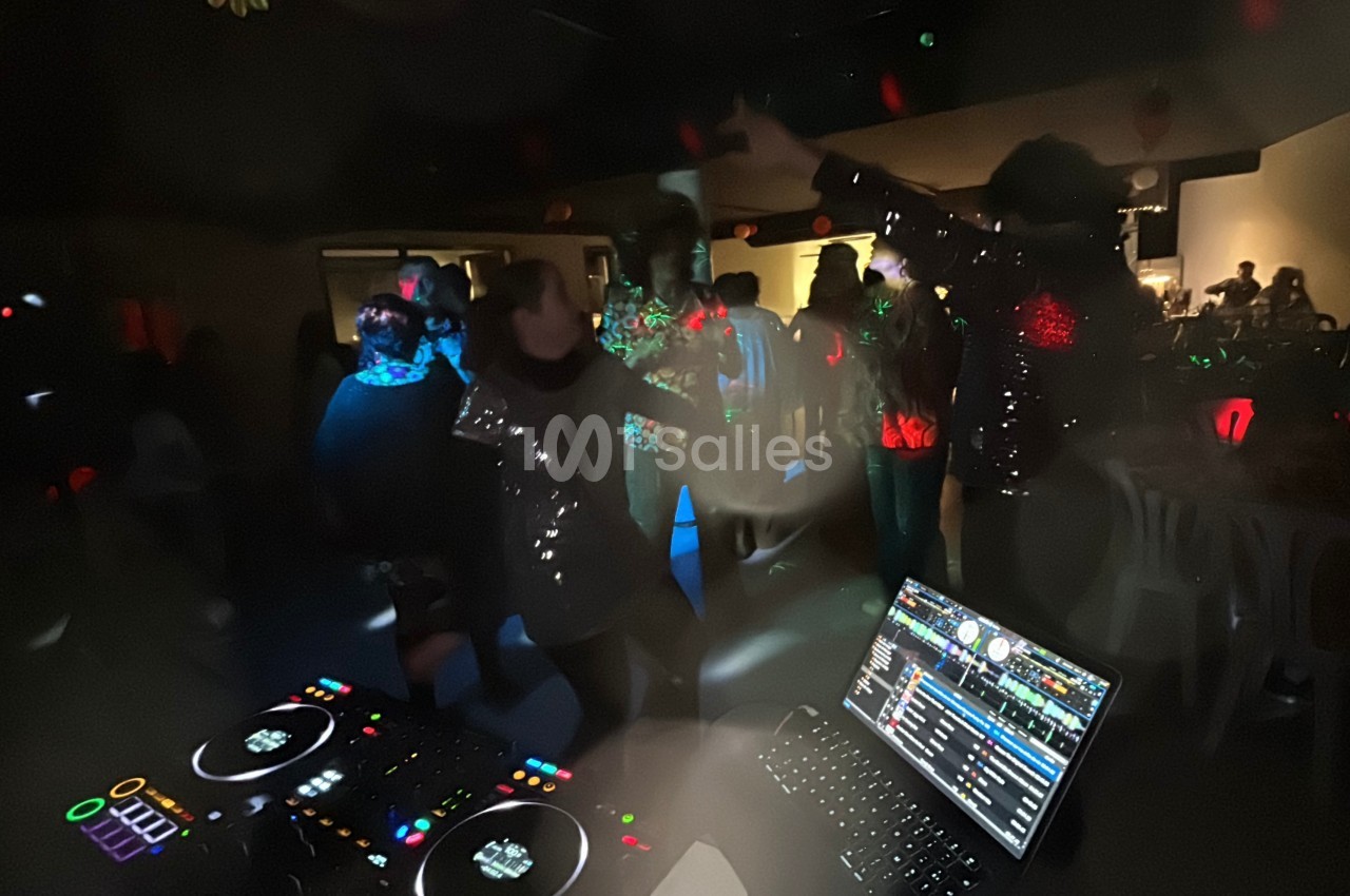 Des personnes dansent dans une ambiance festive et sombre, éclairées par des lumières colorées, avec une table de mixage au…