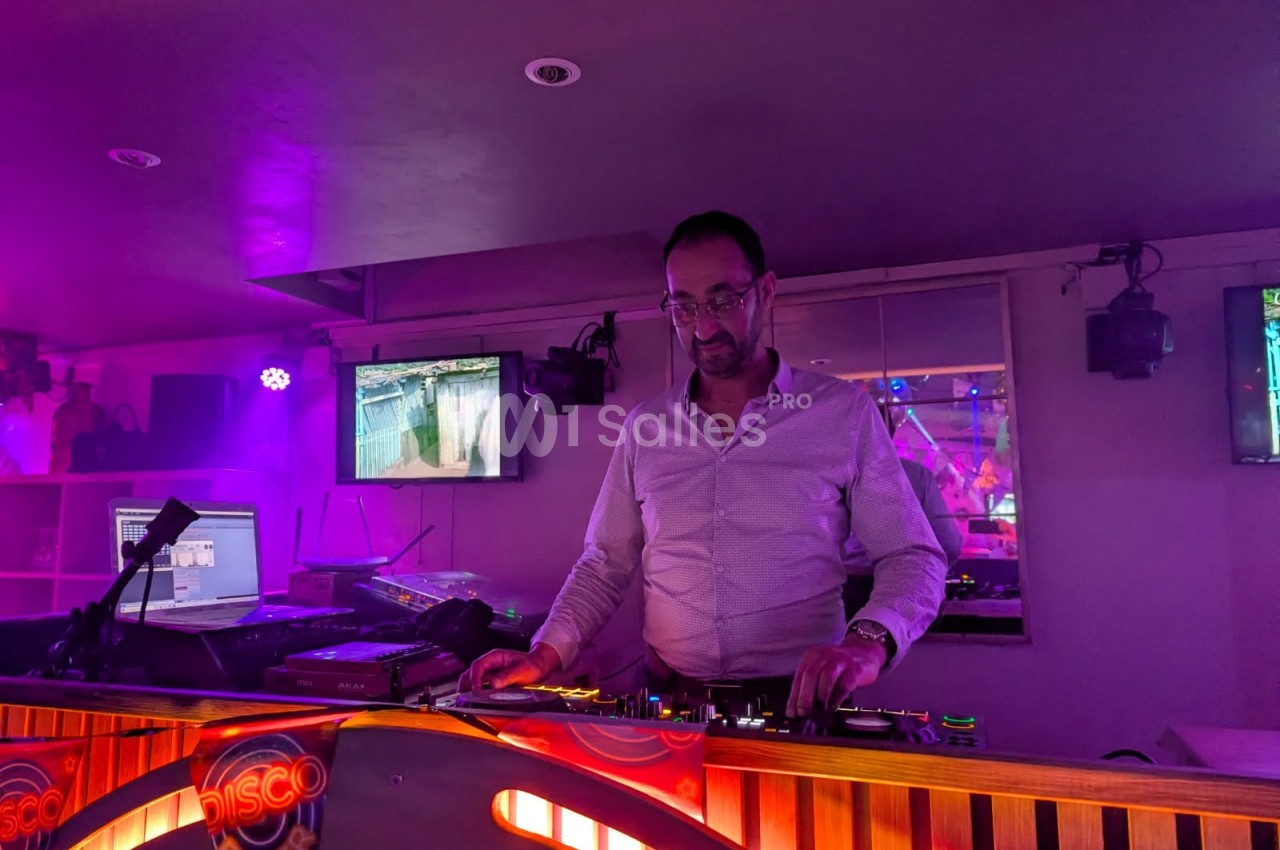 Un DJ mixe de la musique dans un club éclairé par des lumières violettes, avec des écrans en arrière-plan.