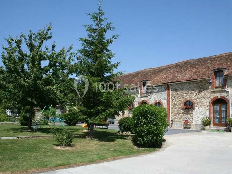 Bâtiment en pierre avec toit en tuiles, entouré d'arbres, pelouse et une balançoire dans un jardin ensoleillé.
