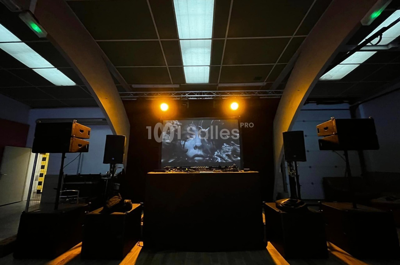 Salle sombre avec une scène équipée de platines DJ, des enceintes et un écran projetant un visage en noir et blanc.