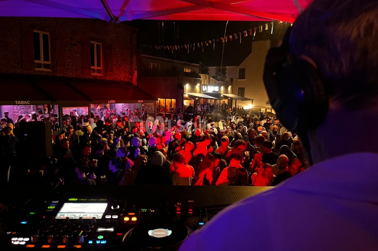 Un DJ mixe sous un chapiteau face à une foule dense lors d'une soirée en plein air, éclairée par des lumières colorées.