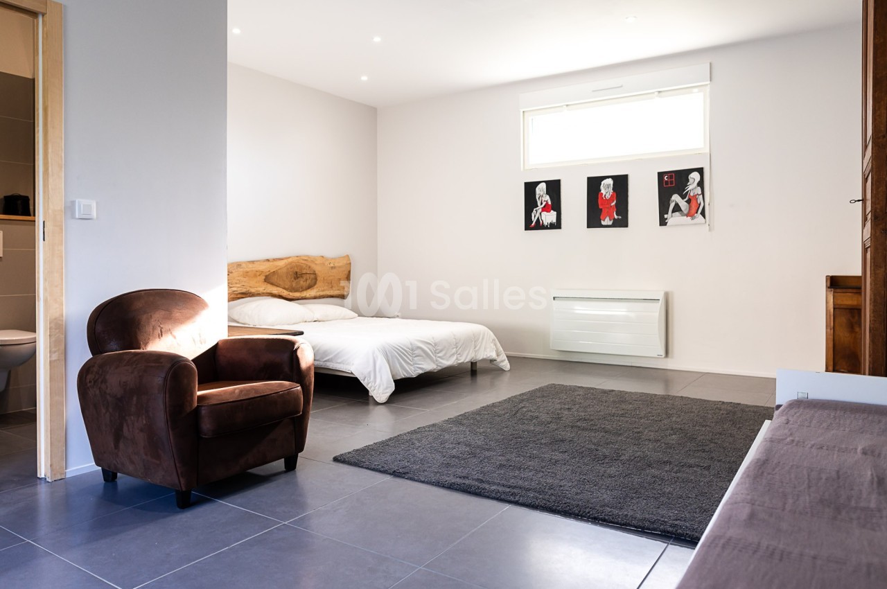 Chambre lumineuse avec lit double, fauteuil en velours marron, tapis gris et décorations murales modernes.
