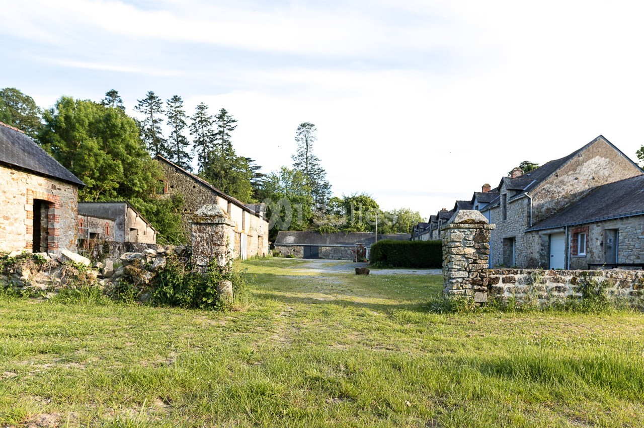 Cour d'une ancienne ferme en pierre avec bâtiments traditionnels, entourée de verdure et d'arbres.