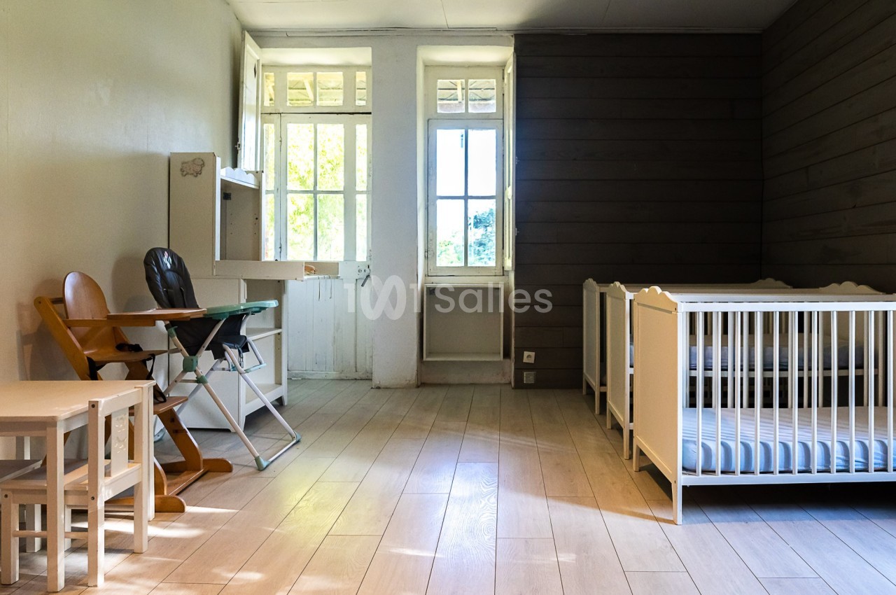Chambre lumineuse avec deux lits pour bébé, une chaise haute, une table et des murs en bois et en blanc.