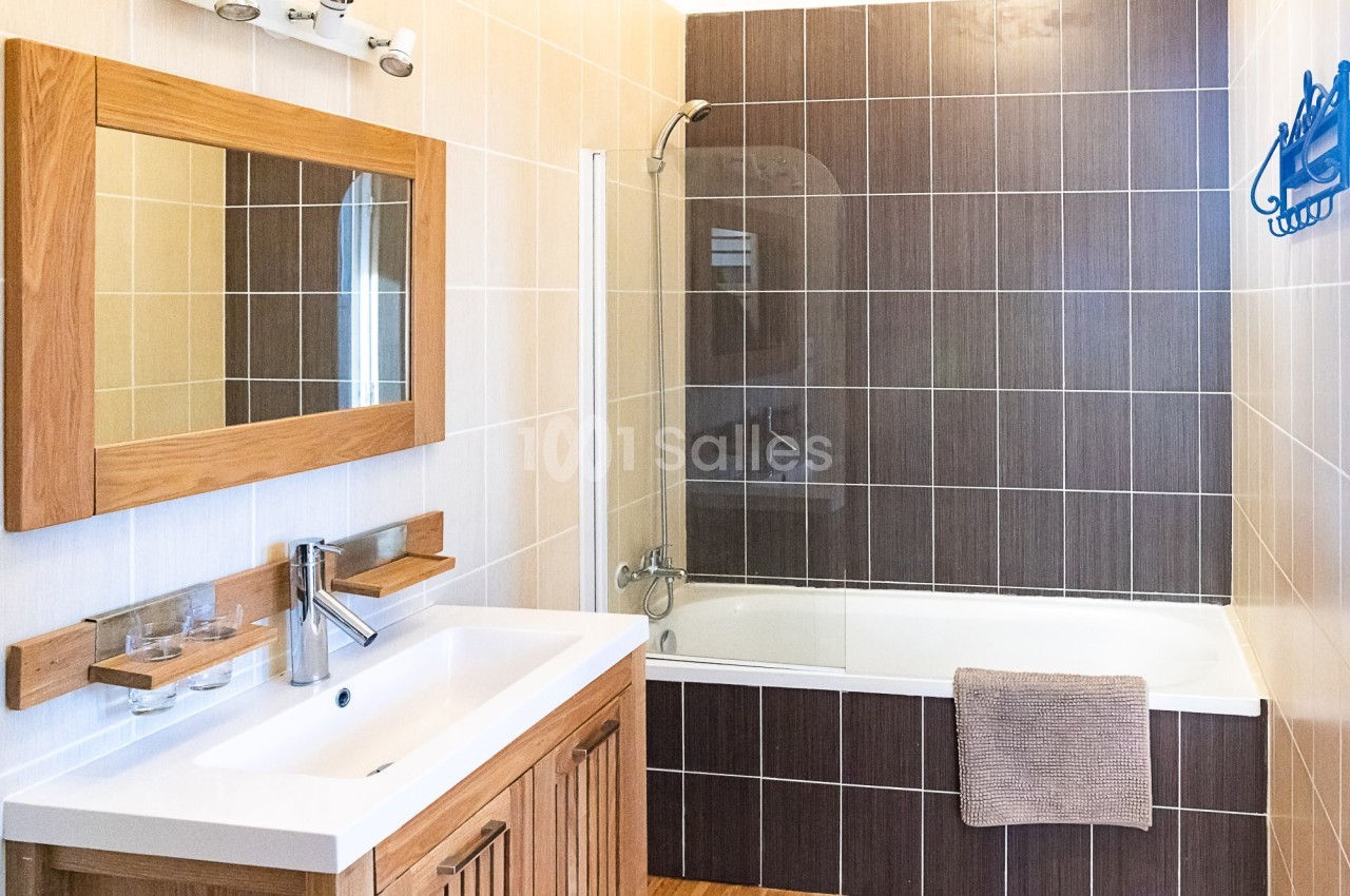 Salle de bain avec lavabo en bois, miroir assorti, baignoire avec paroi vitrée et carrelage beige et marron.