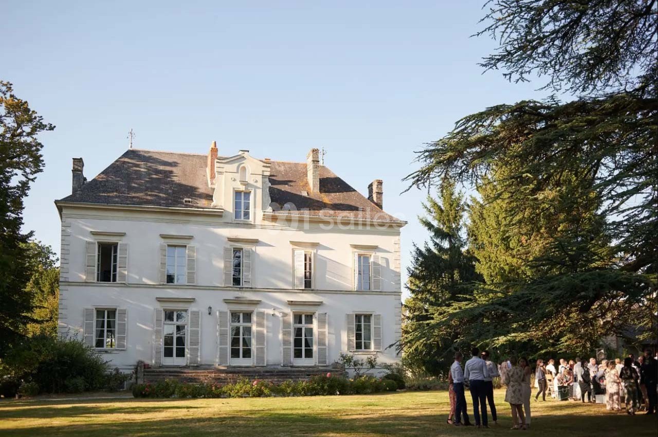 Façade d'un manoir blanc entouré d'un jardin, avec un groupe de personnes rassemblées à l'ombre des arbres.