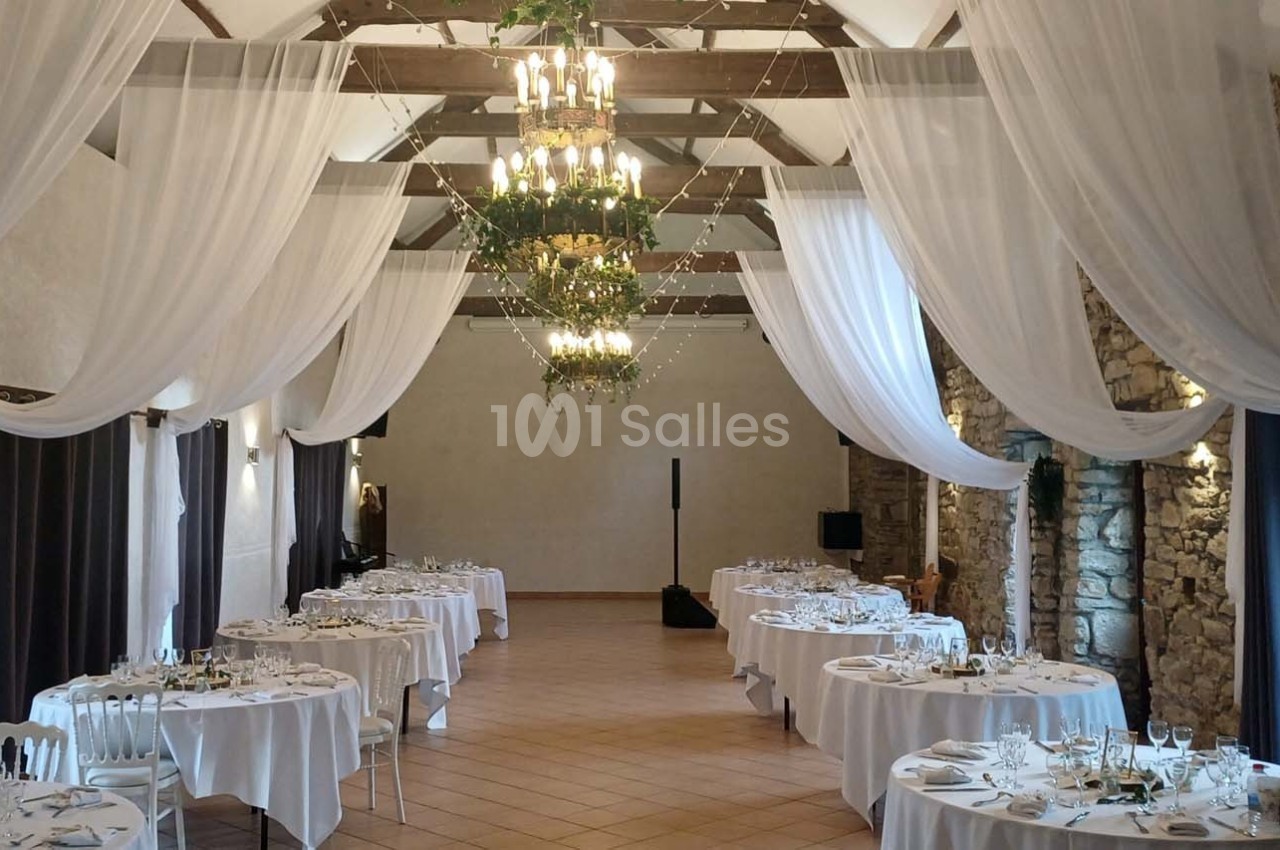 Salle de réception avec tables rondes dressées, rideaux blancs suspendus et murs en pierre apparente.