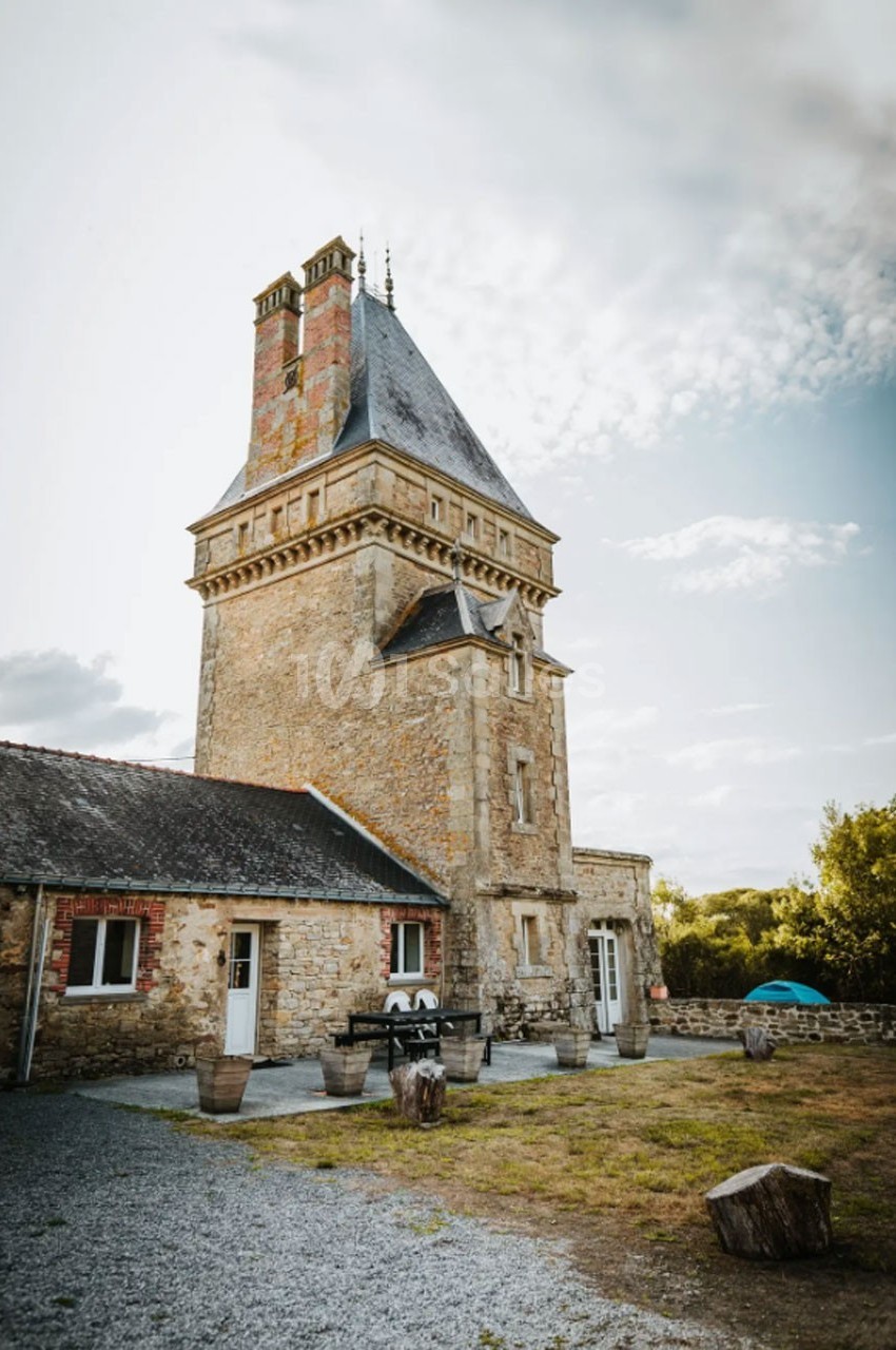 Tour en pierre avec toit en ardoise, attenante à une maison, entourée d'un jardin et d'une tente visible au loin.