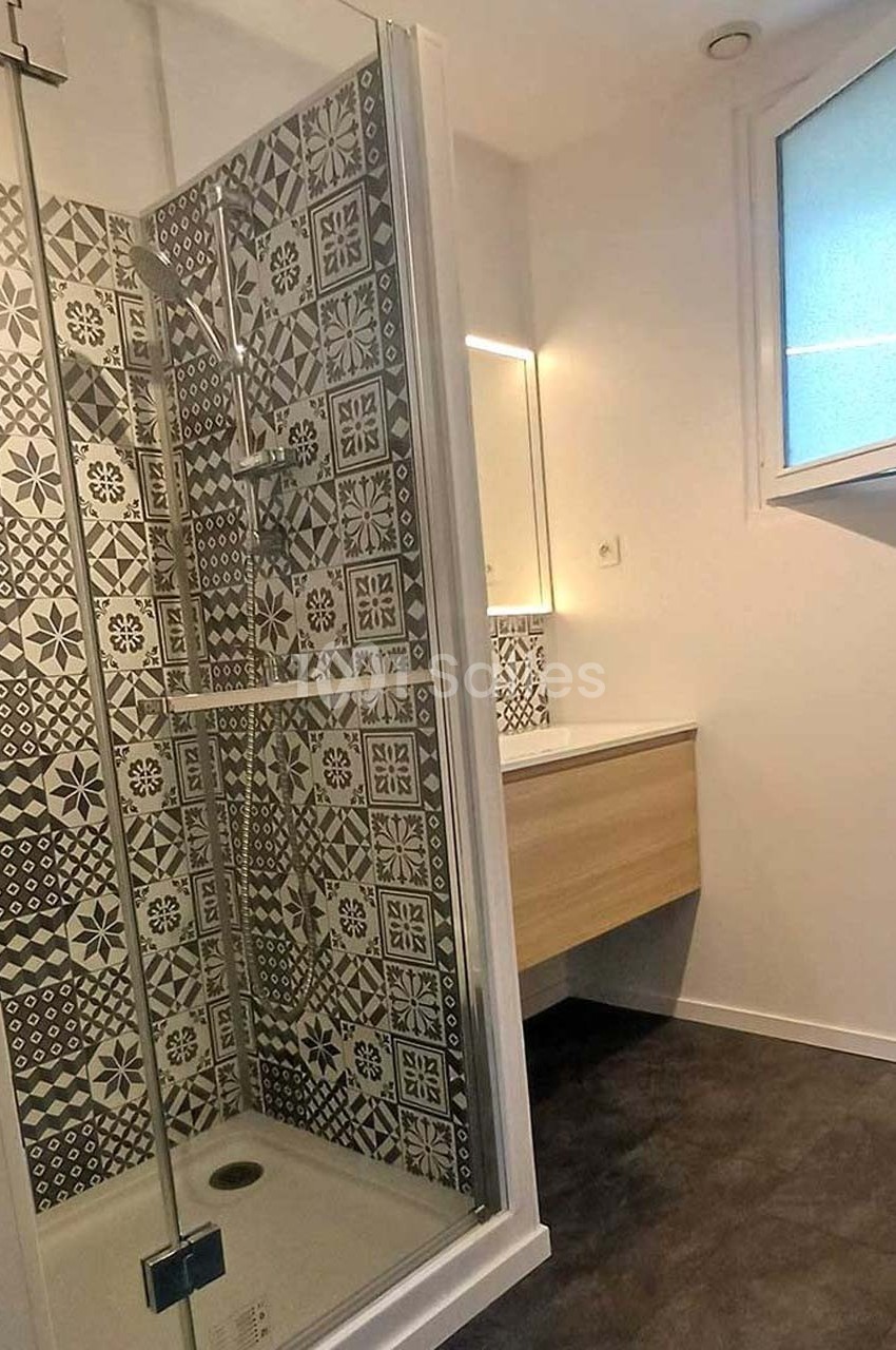 Douche avec paroi vitrée, carrelage à motifs géométriques noirs et blancs, meuble vasque et fenêtre en arrière-plan.