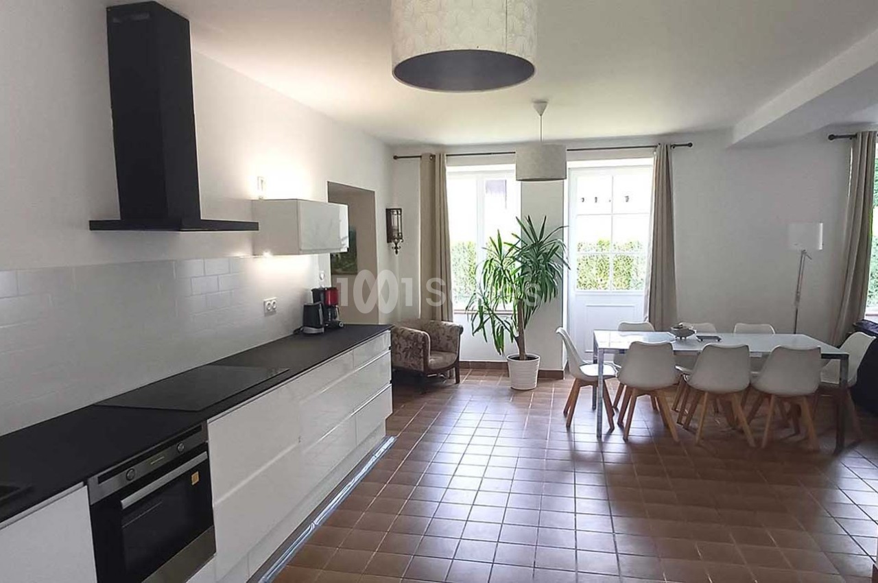 Cuisine ouverte moderne avec îlot, coin repas, table blanche et chaises, éclairée par de grandes fenêtres.