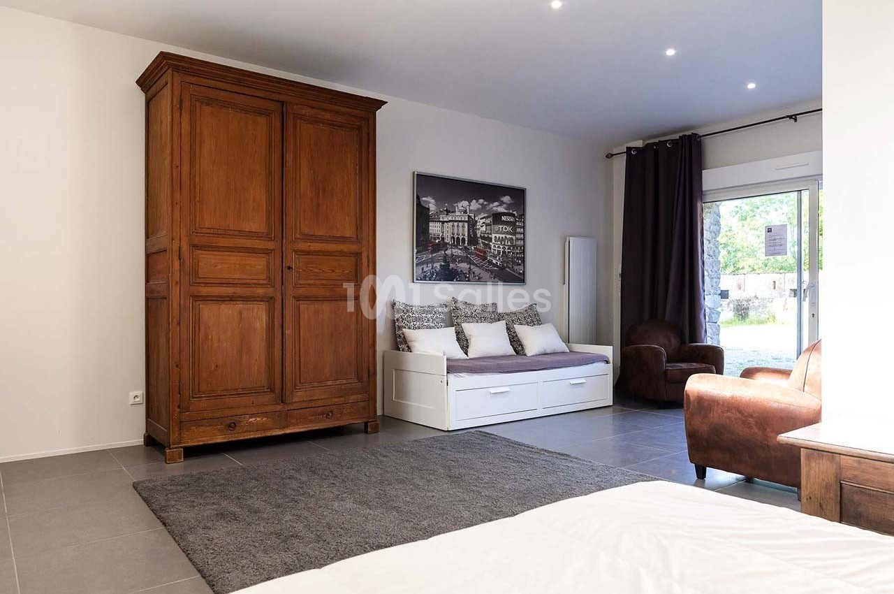 Chambre lumineuse avec armoire en bois, canapé-lit, fauteuil en cuir, tapis gris et grande fenêtre donnant sur l'extérieur.