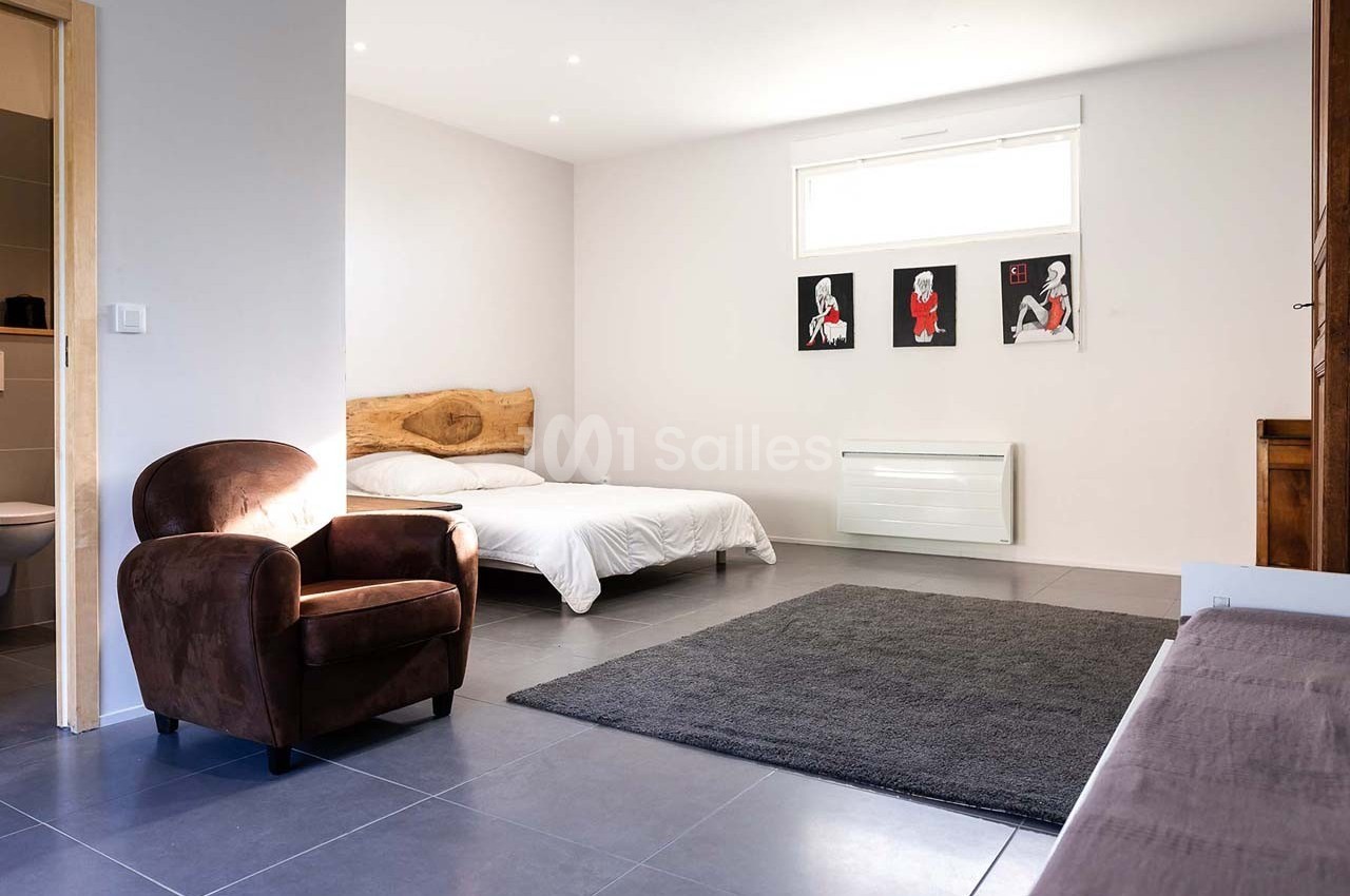 Chambre lumineuse avec lit double, fauteuil en tissu marron, tapis gris et décorations murales modernes.