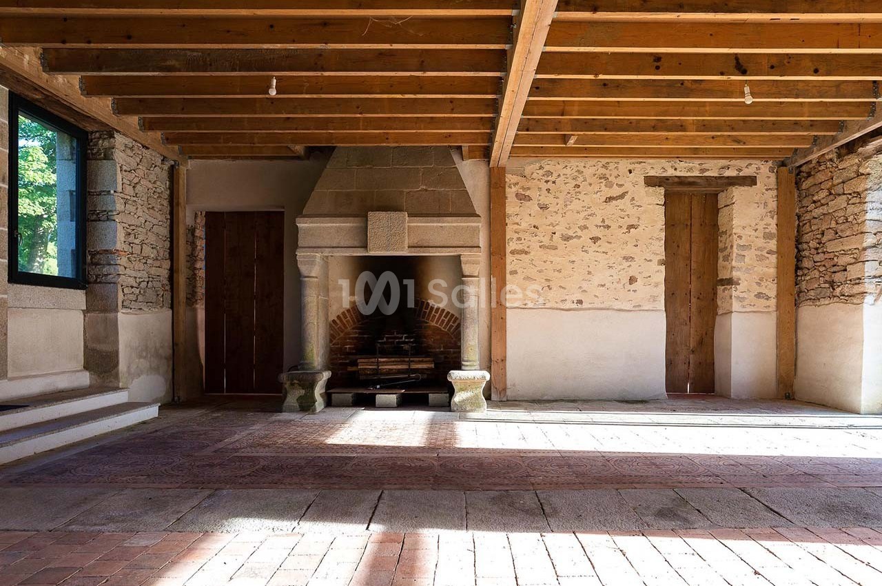 Salle avec murs en pierre, cheminée centrale, plafond en poutres apparentes et sol en briques.